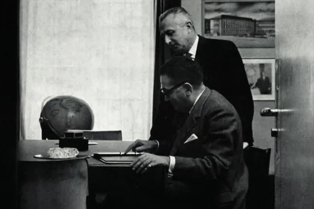 Raoul Perret consulte des documents dans un bureau Universal Genève en noir et blanc avec globe et secrétaire.